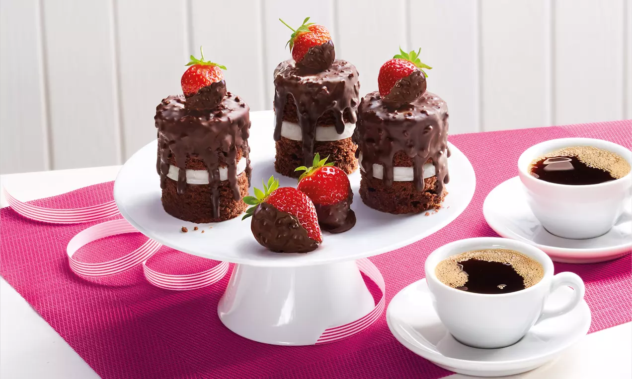 Schokotörtchen in zylindrischer Form mit dunkler Knusperglasur, gefüllt mit heller Creme, dekoriert mit Erdbeeren auf weißer Kuchenplatte