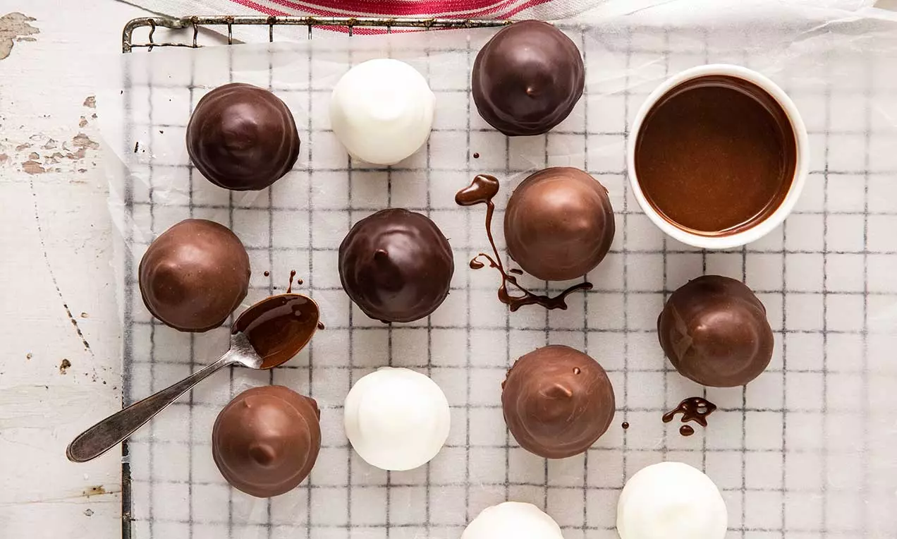 Petits dômes chocolatés lisses et brillants, bruns ou blancs, posés sur une grille avec une cuillère et un bol de sauce