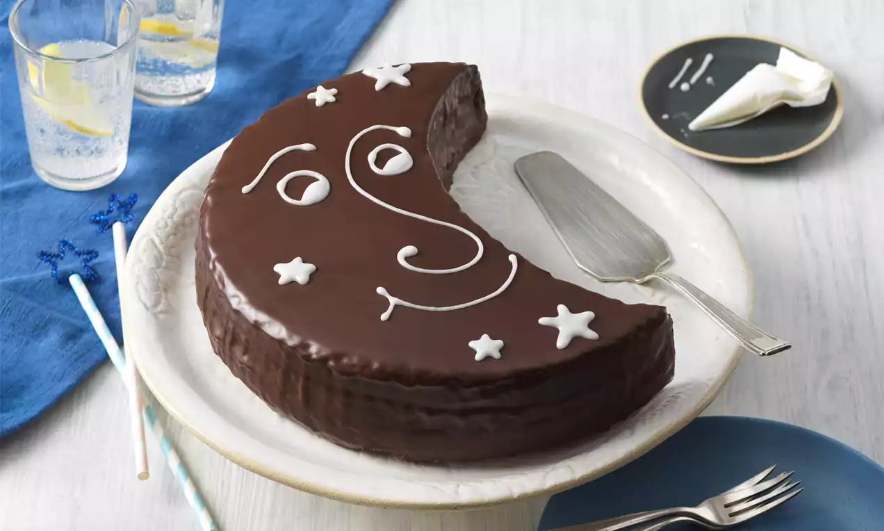 Gâteau en croissant brun chocolat brillant décoré de motifs blancs en lune et étoiles sur une assiette avec des couverts et nappe bleue