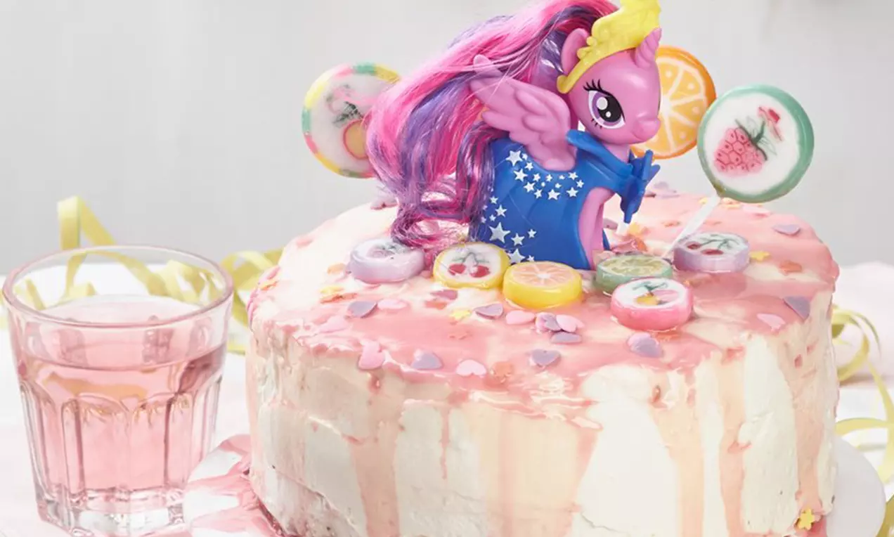 Gâteau rond recouvert de glaçage rose avec des bonbons colorés et une figurine Petit Poney entourée de confettis en sucre