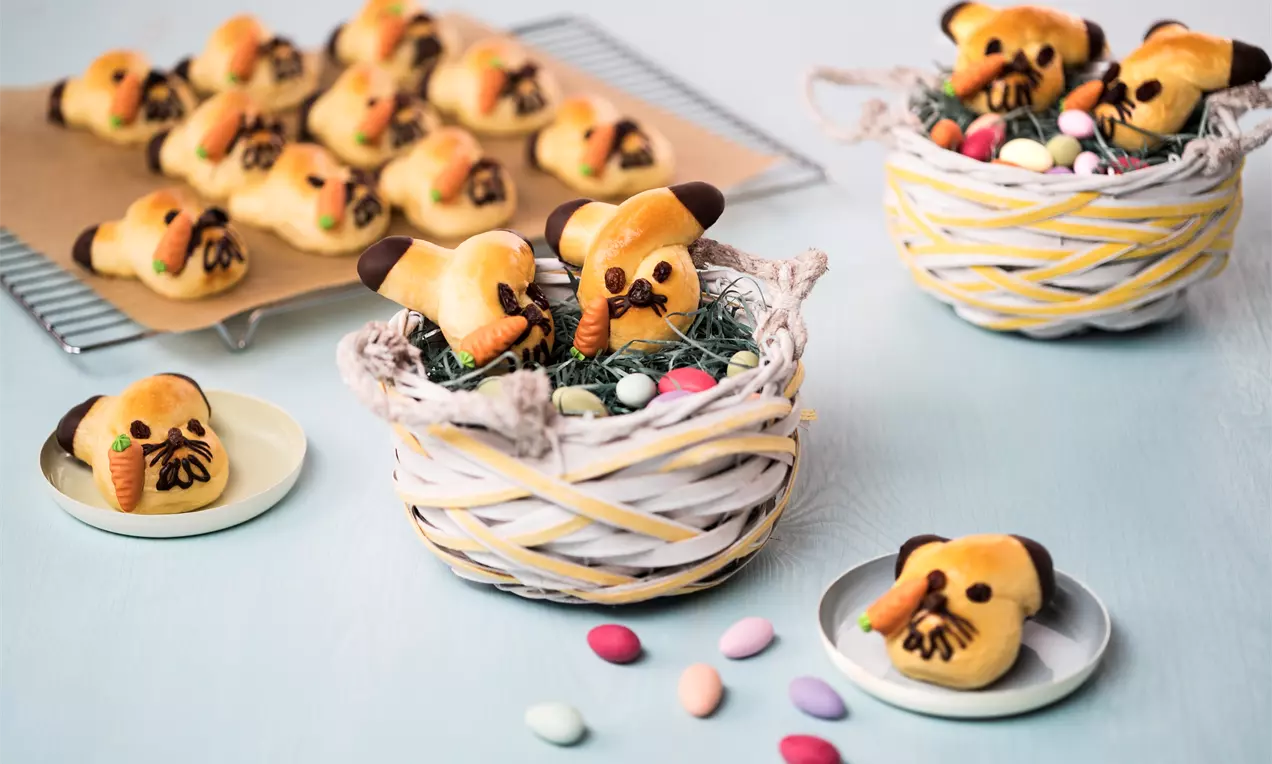Goldbraune Häschengebäcke mit Schoko-Dekor und Mandeln auf kunstvoll geflochtenen Körbchen mit grünem Ostergras und bunten Dragees