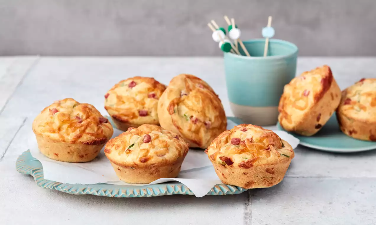 Herzhafte Muffins goldgelb und locker, mit fein sichtbaren Zutaten, auf einem türkisfarbenen Teller mit Papier, daneben ein Becher mit Spießen