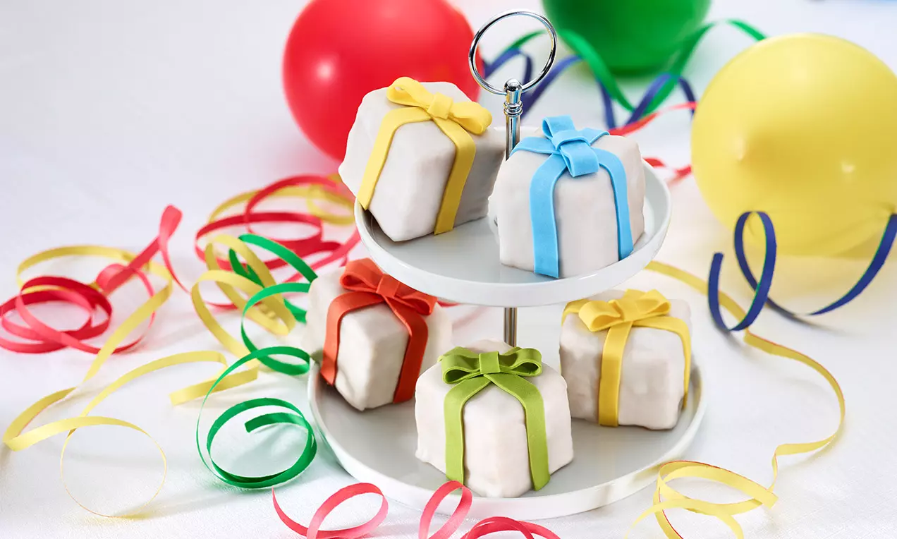 Weiße würfelförmige Pralinen mit farbigen Zuckerguss-Schleifen auf einer silbernen Etagere, dekoriert mit Luftschlangen und bunten Ballons