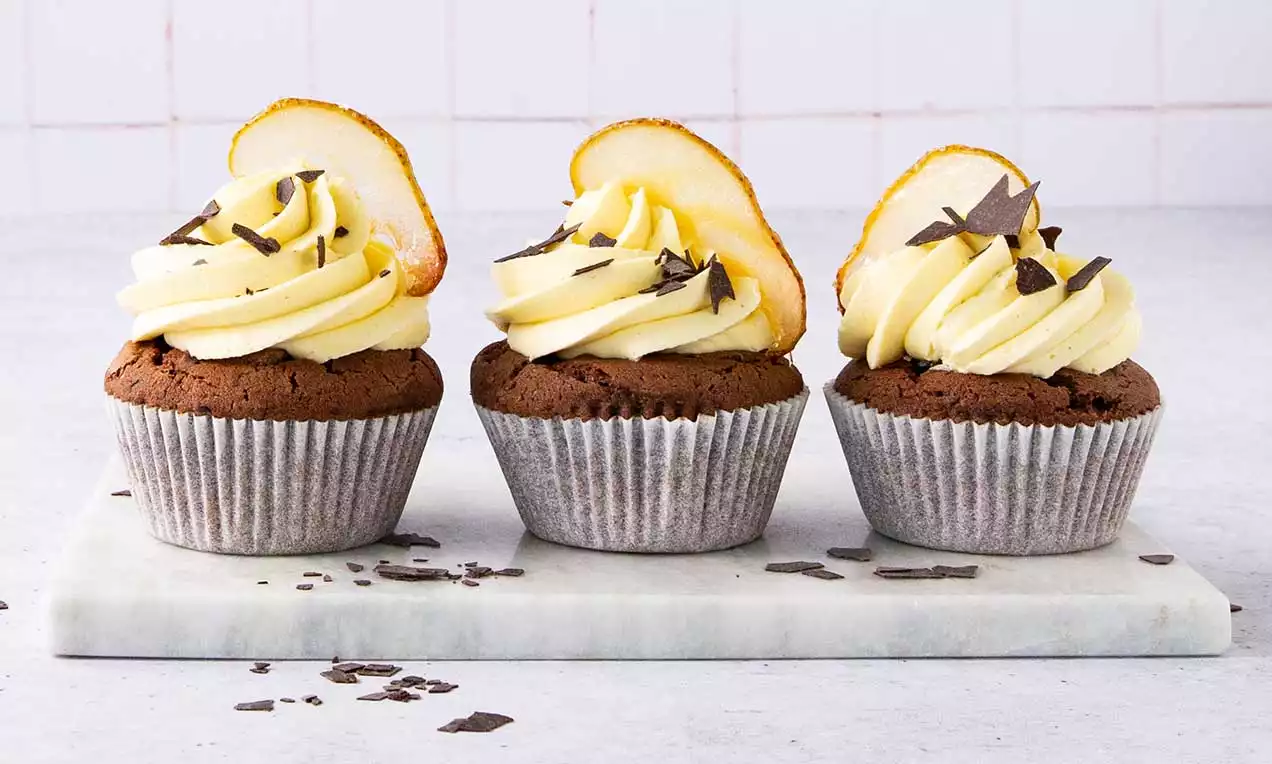 Dunkelbraune Cupcakes mit geriffelter Papierhülle, bestrichen mit gelber Sahnecreme, Birnenscheiben und dunklen Schokoraspeln auf Marmorplatte