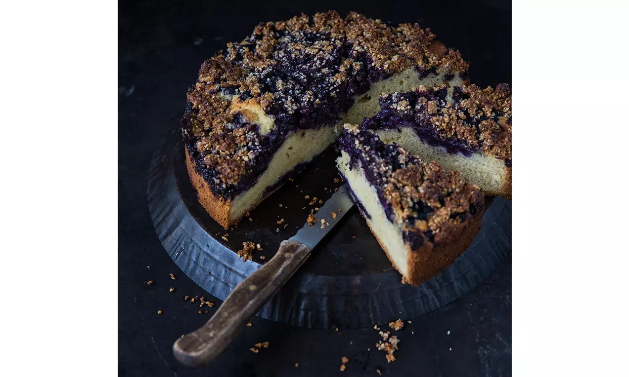 Gâteau rond moelleux avec une couche de myrtilles foncées, surmonté de crumble doré, posé sur une assiette noire avec un couteau
