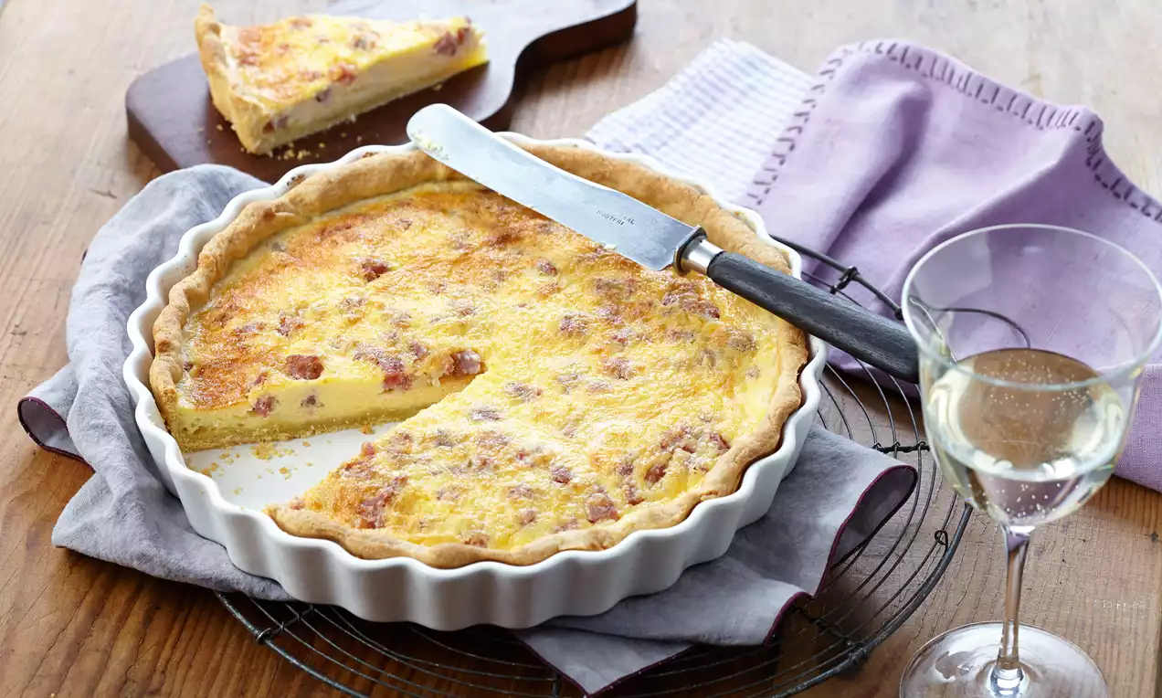Quiche Lorraine mit goldgelber Füllung und röschem Rand in einer weißen Form auf einem violetten Tuch, daneben Glas und Kuchenstück