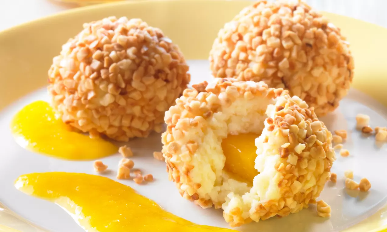 Knödel dorati ricoperti di granella croccante, con cuore di cremosa pesca gialla su un piatto chiaro decorato con crema di frutta nello stesso tono