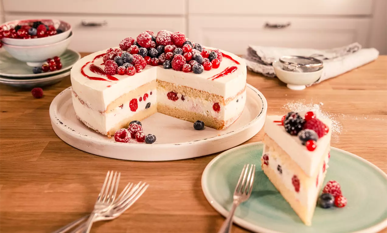 Torta soffice a strati con crema chiara e frutti di bosco rossi e blu, decorata con zucchero a velo e coulis di frutti rossi su un piatto bianco