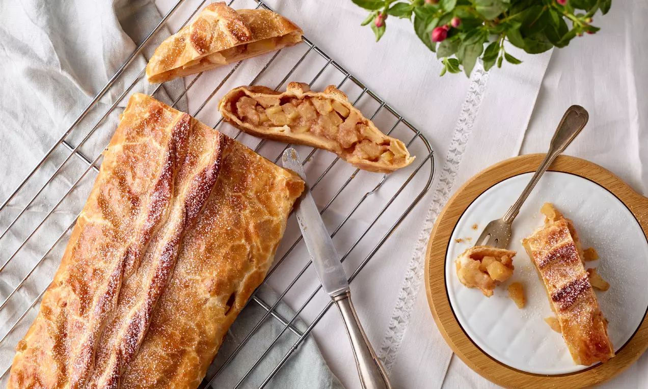 Apfelstrudel doré en pâte feuilletée délicatement sucrée, garni de pommes fondantes, tranché sur une grille avec une assiette en bois et couverts