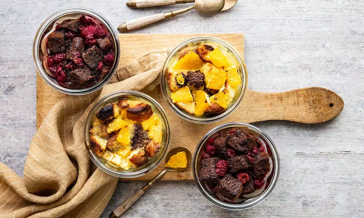 Gratin dolce in bicchieri di vetro con strati colorati di crema chiara, frutti rossi vivaci e pezzi dorati di pane su un tagliere di legno