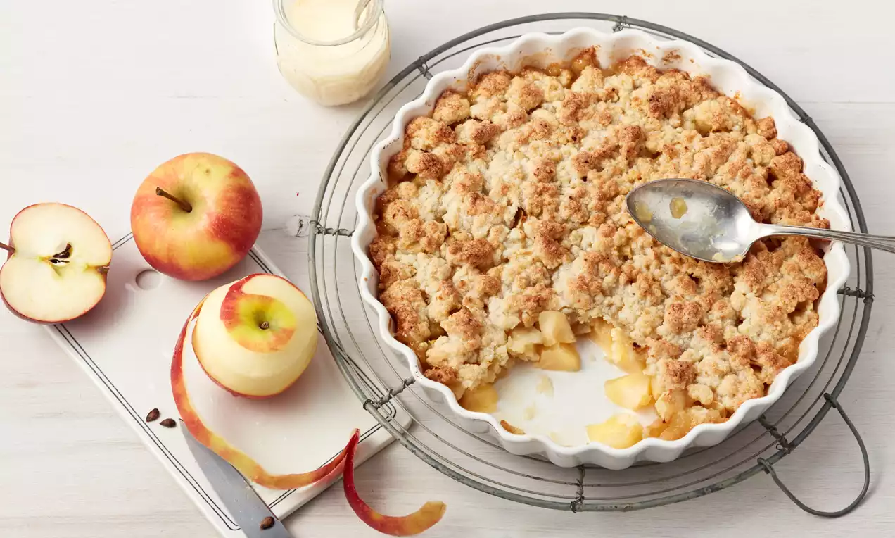 Goldbrauner Crumble mit grobkrümeliger Oberfläche in weißer runder Form, daneben frische Äpfel, Apfelschalen und eine Serviergabel