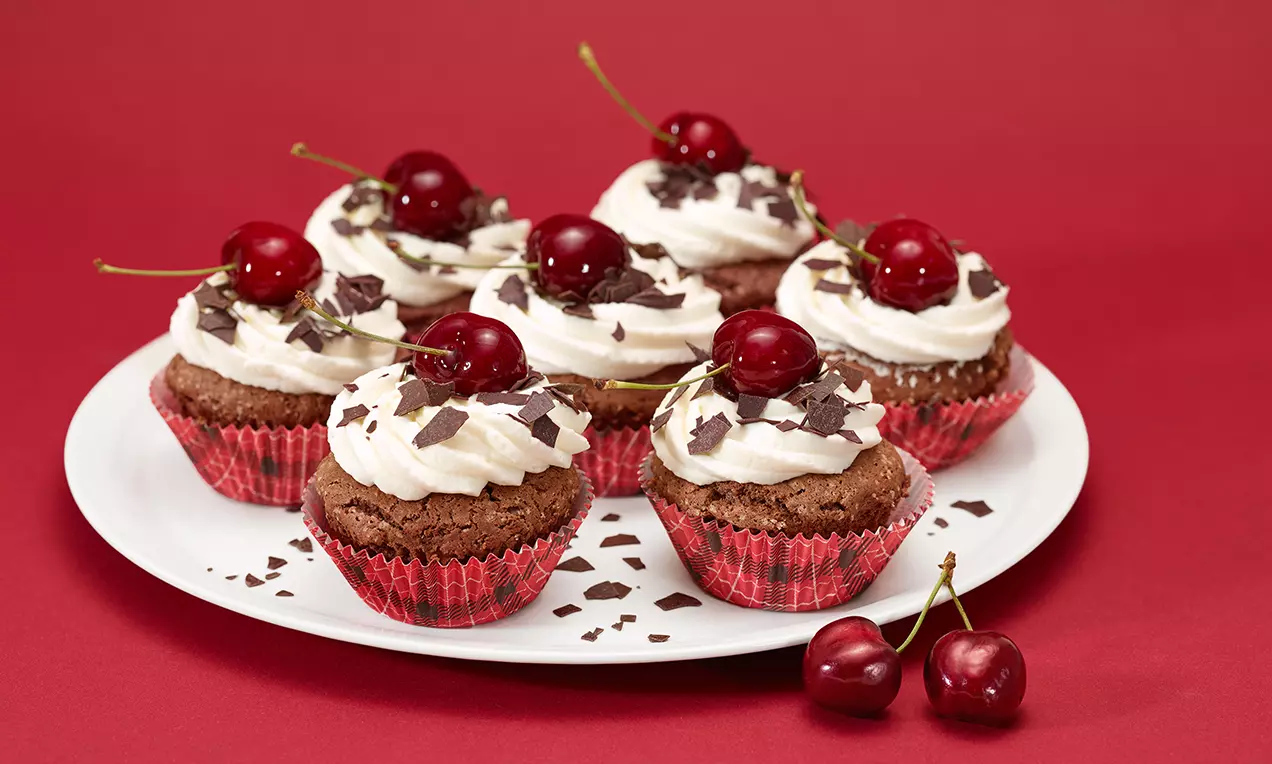 Cupcake al cioccolato in pirottini rossi, con ciuffi di panna bianca, scaglie di cioccolato fondente e ciliegie lucide su un piatto bianco