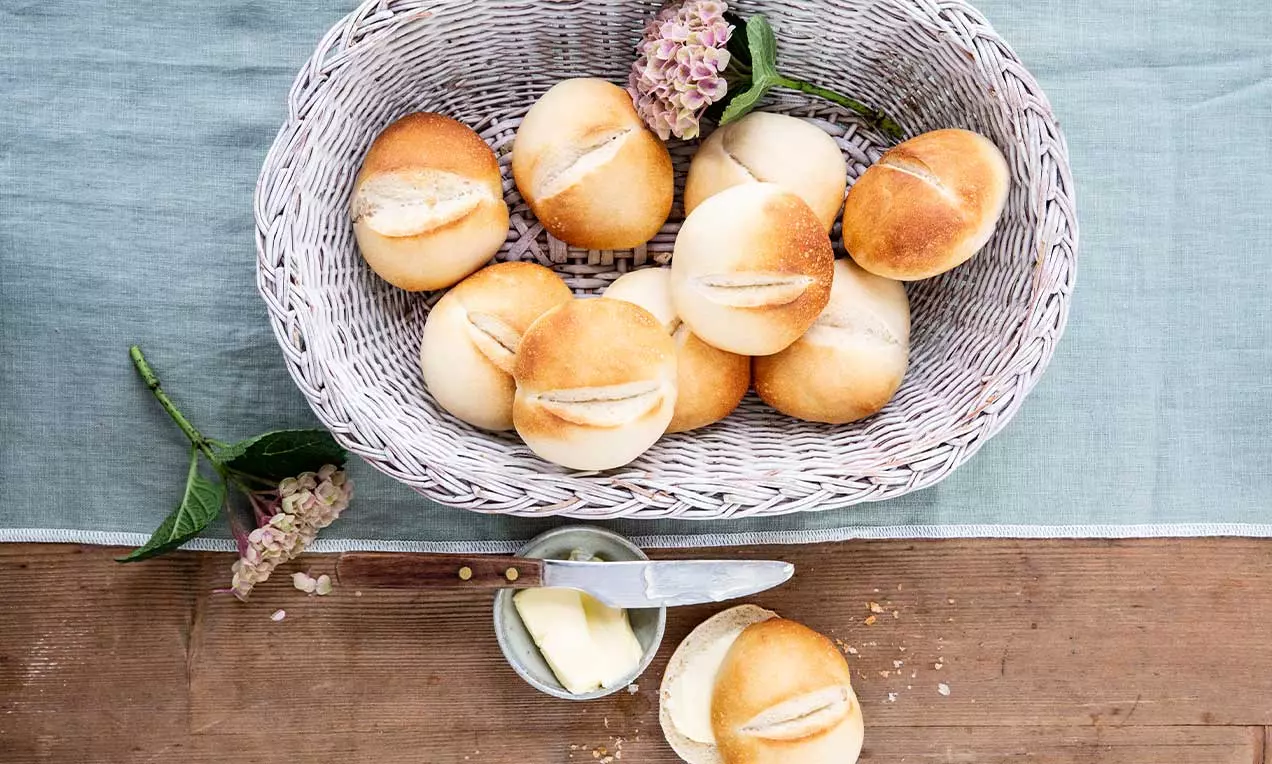 Kleine, goldbraun gebackene Brötchen mit feiner Rille in weidenweißer Korbschale auf blauem Stoff neben Butter, Messer und Holz