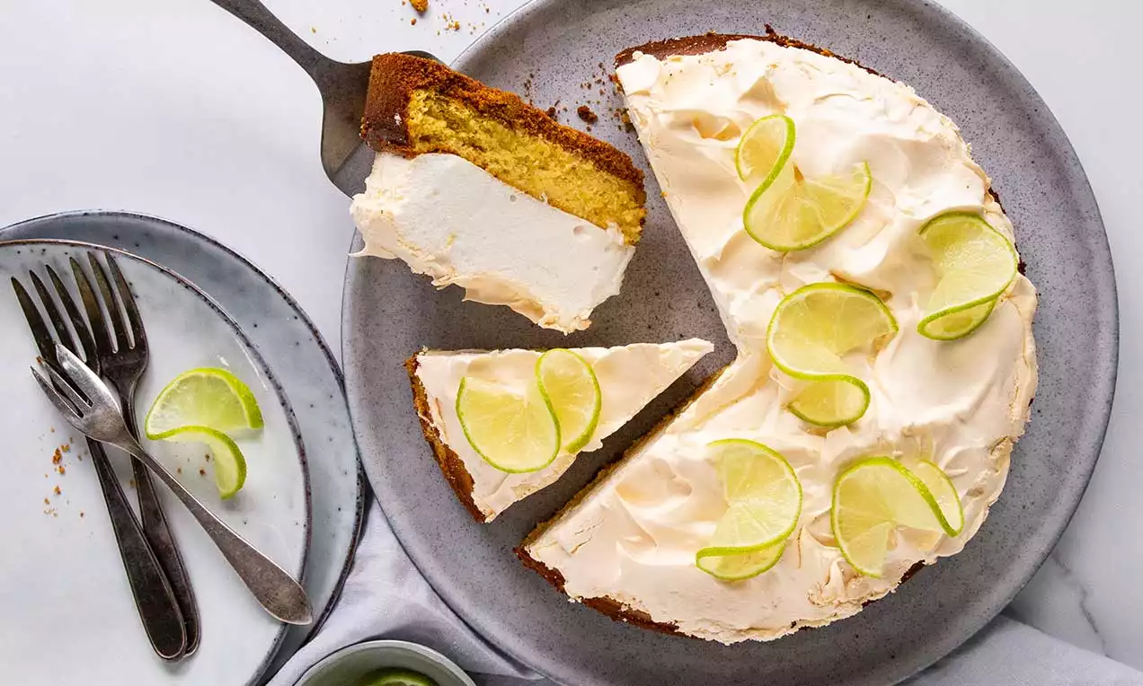 Limetten-Tarte mit heller Meringue-Haube, belegt mit frischen Limettenscheiben, serviert auf grauem Teller und angeschnitten mit Kuchenheber