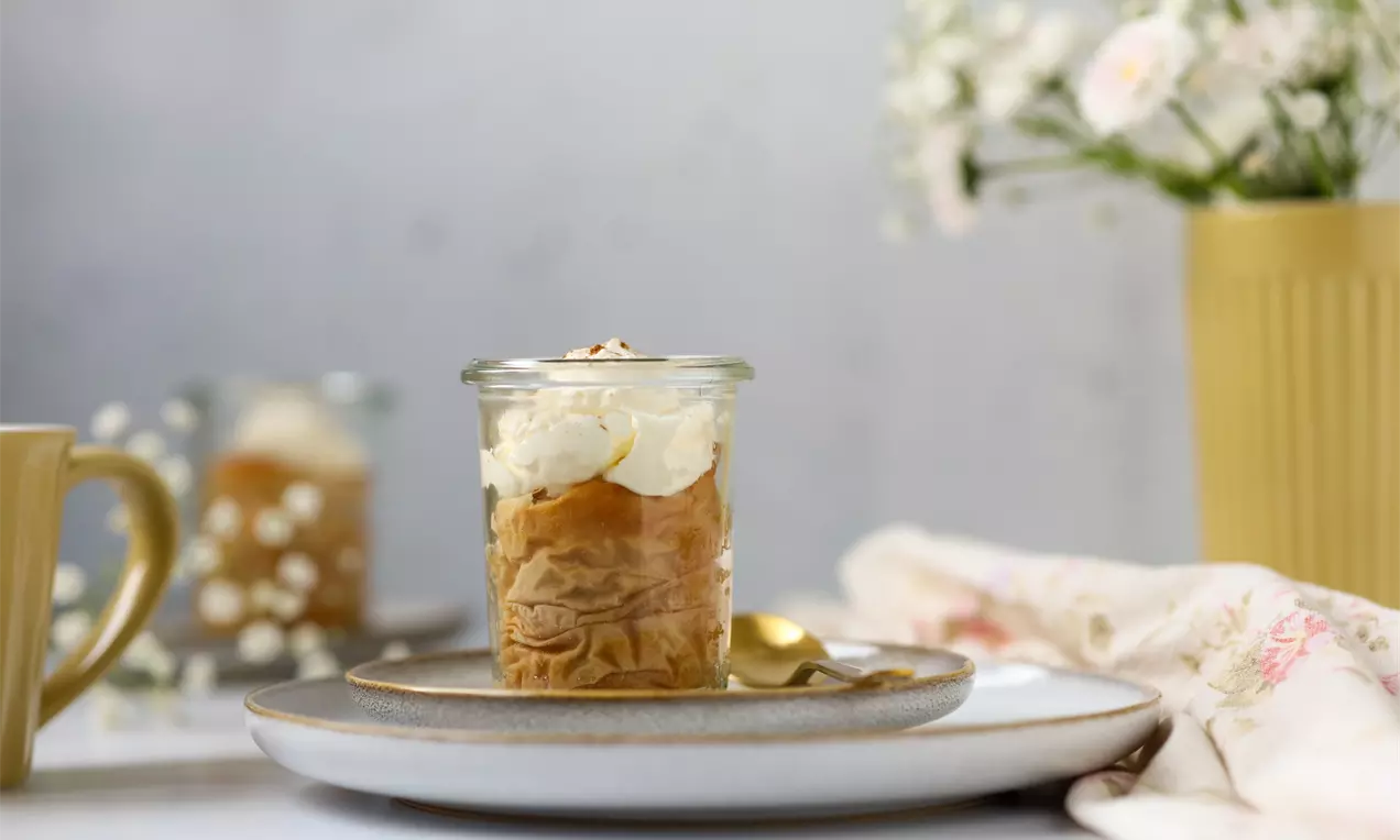 Apfelstrudel im klaren Glas mit goldbraun geschichteter Füllung, Sahnehaube und Zimt bestäubt auf grauem Teller mit goldener Löffeldekoration