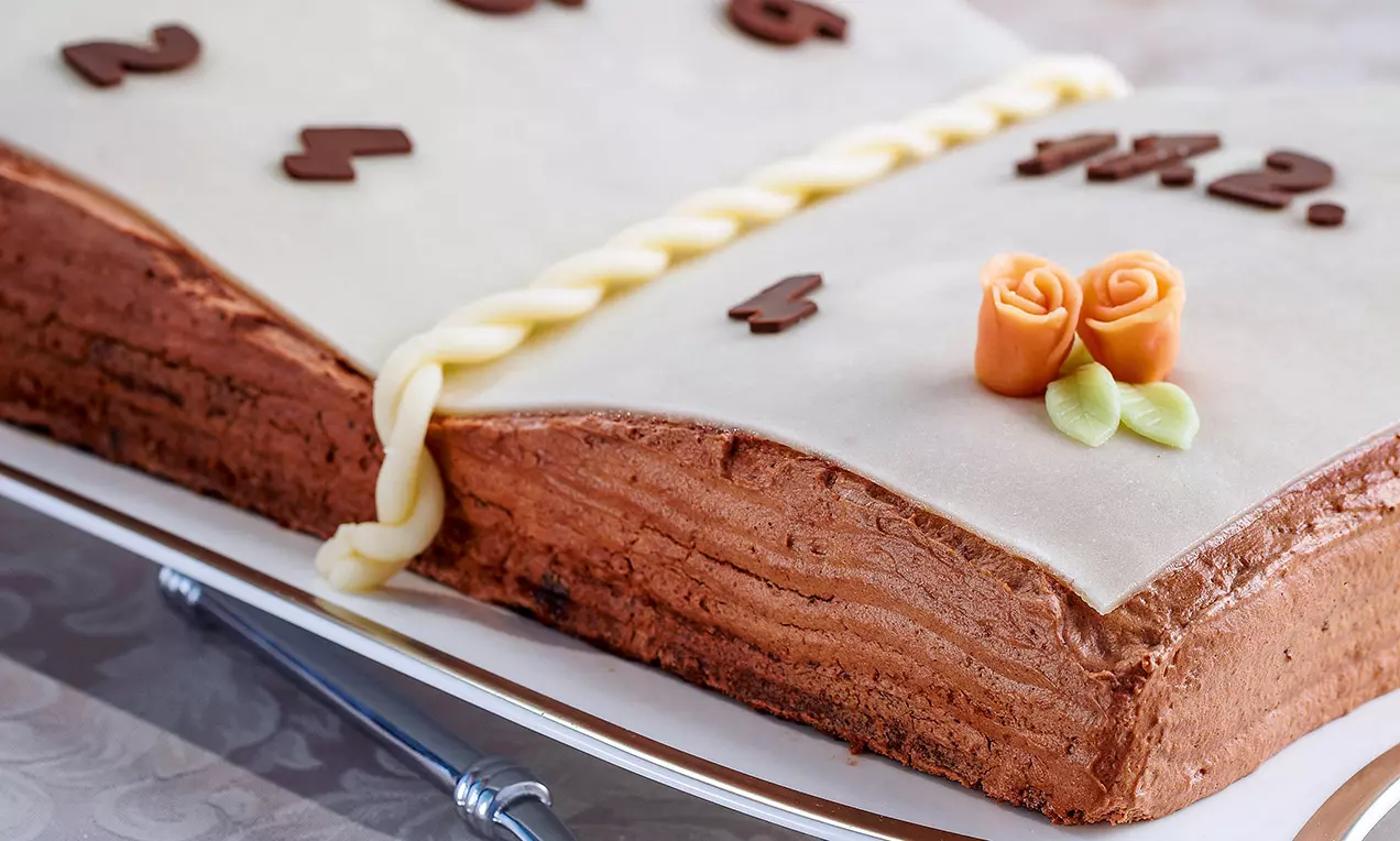 Gâteau en forme de livre ouvert avec glaçage crème beige, bord tressé, décoré de roses orange et feuilles vertes sur une assiette blanche