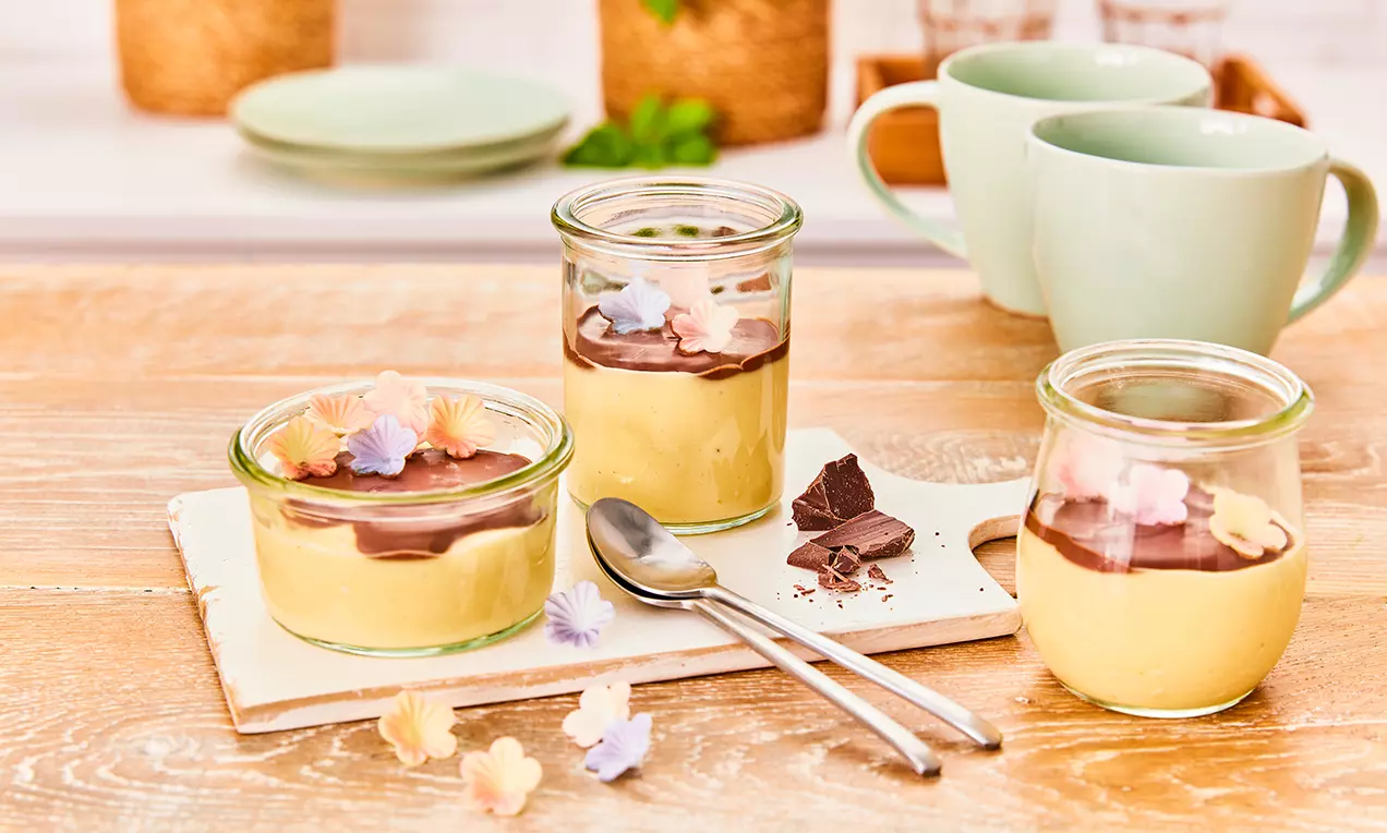 Veganer Vanillepudding in kleinen Gläsern mit cremig-glatter Schokosoße, dekoriert mit zarten essbaren Blüten auf Holzbrett serviert