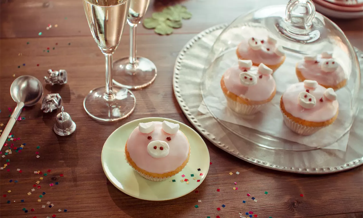 Rosa Cupcake mit Schweinegesicht-Dekor, Ohren und Nase aus Zuckerguss, auf grünem Teller mit bunten Konfettistreuseln und Sektgläsern