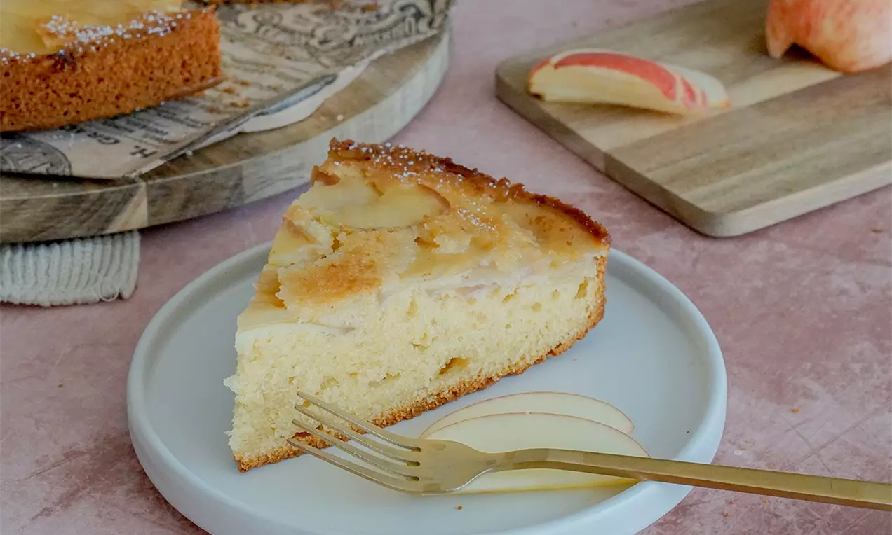 Ein goldbraunes Stück Upside-Down-Apfelkuchen mit karamellisierten Apfelscheiben auf einem weißen Teller mit goldener Gabel und Apfelstücken