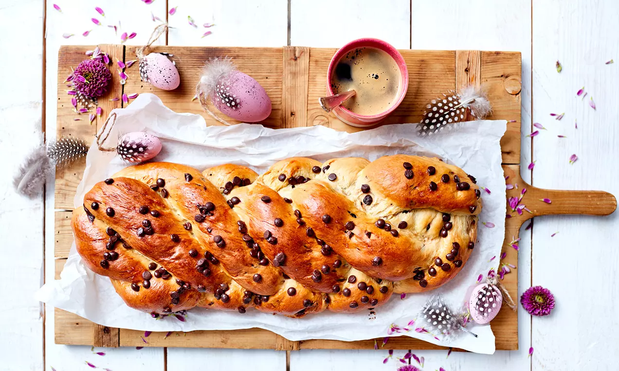 Fünfsträngiger Zopf aus glänzend goldbraunem Hefeteig mit Schokoladenstücken, auf Backpapier auf einem Holzbrett mit Kaffeetasse und Osterdeko