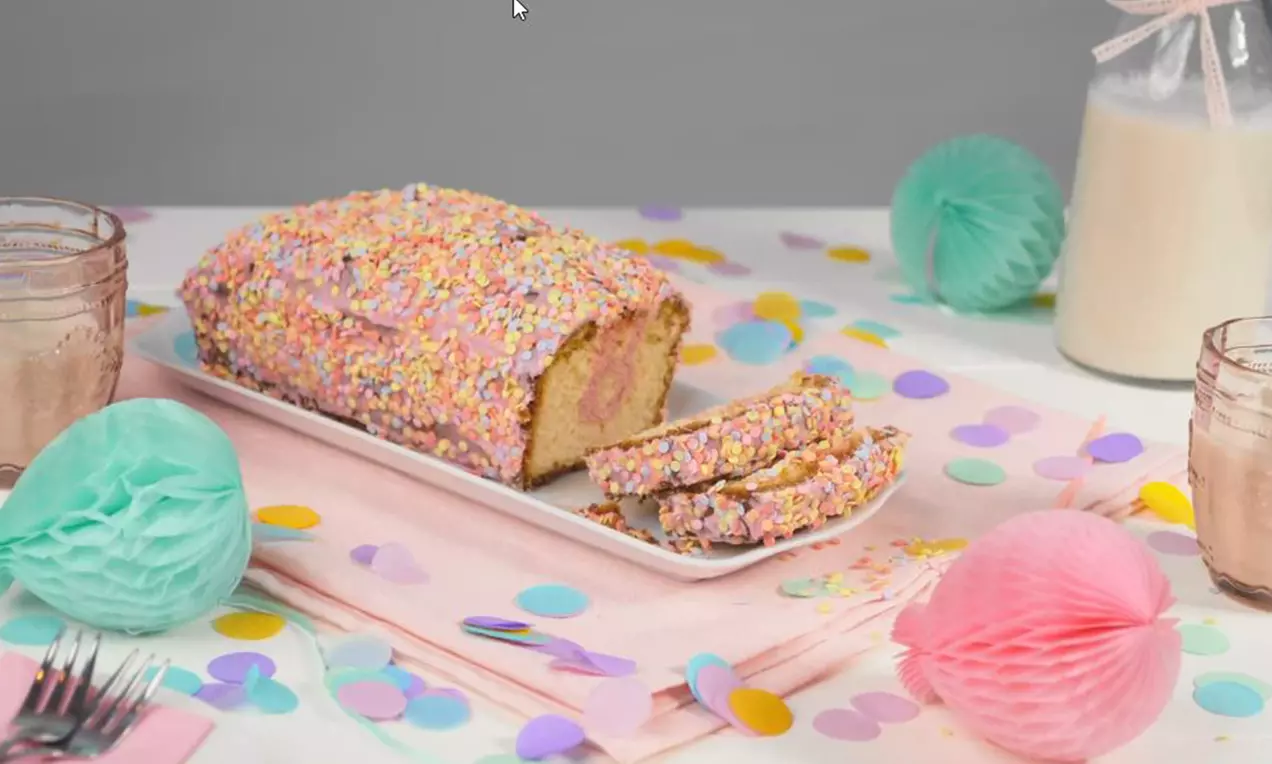 Überraschungskuchen mit gelber Krume und rosa Füllung, umhüllt von bunten Streuseln, auf weißer Platte mit pastellfarbener Tischdeko