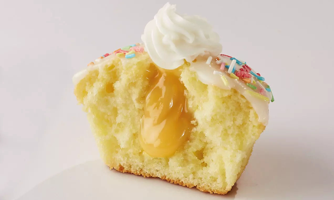 Cupcake doré à la mie moelleuse, garni de glaçage blanc, décoré de perles colorées et rempli d'un cœur fondant de citron jaune lumineux