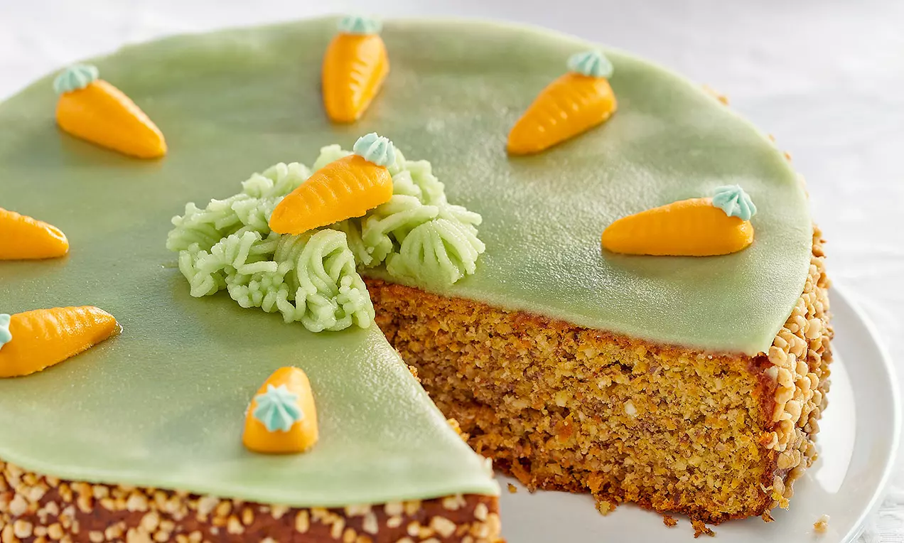 Gâteau aux carottes à la texture dense avec glaçage vert tendre garni de petites carottes en pâte d’amande et bordé de noisettes hachées