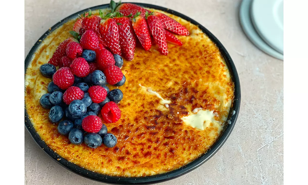 Creme-Brûlée-Tarte mit goldbrauner Karamellkruste, frischen Erdbeeren, Himbeeren und Blaubeeren auf einem schwarzen Teller