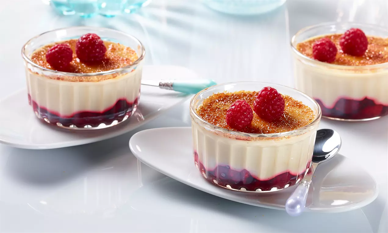 Crème Brûlée mit karamellisierter goldbrauner Kruste, roten Himbeeren und tiefroter Fruchtschicht in Glasförmchen auf weißen Tellern