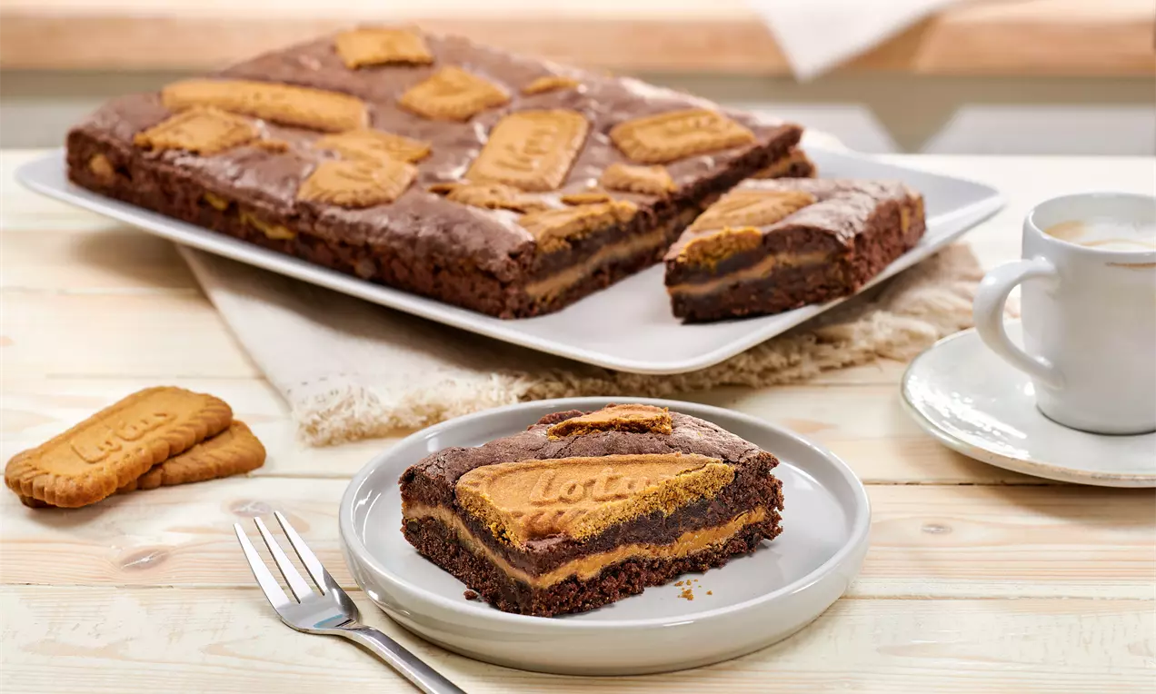 Rechteckiger Lotus-Brownie mit schokoladiger Textur, Biscoff-Keksen belegt, serviert auf weißem Teller, ergänzt durch Gabel und Kaffeetasse