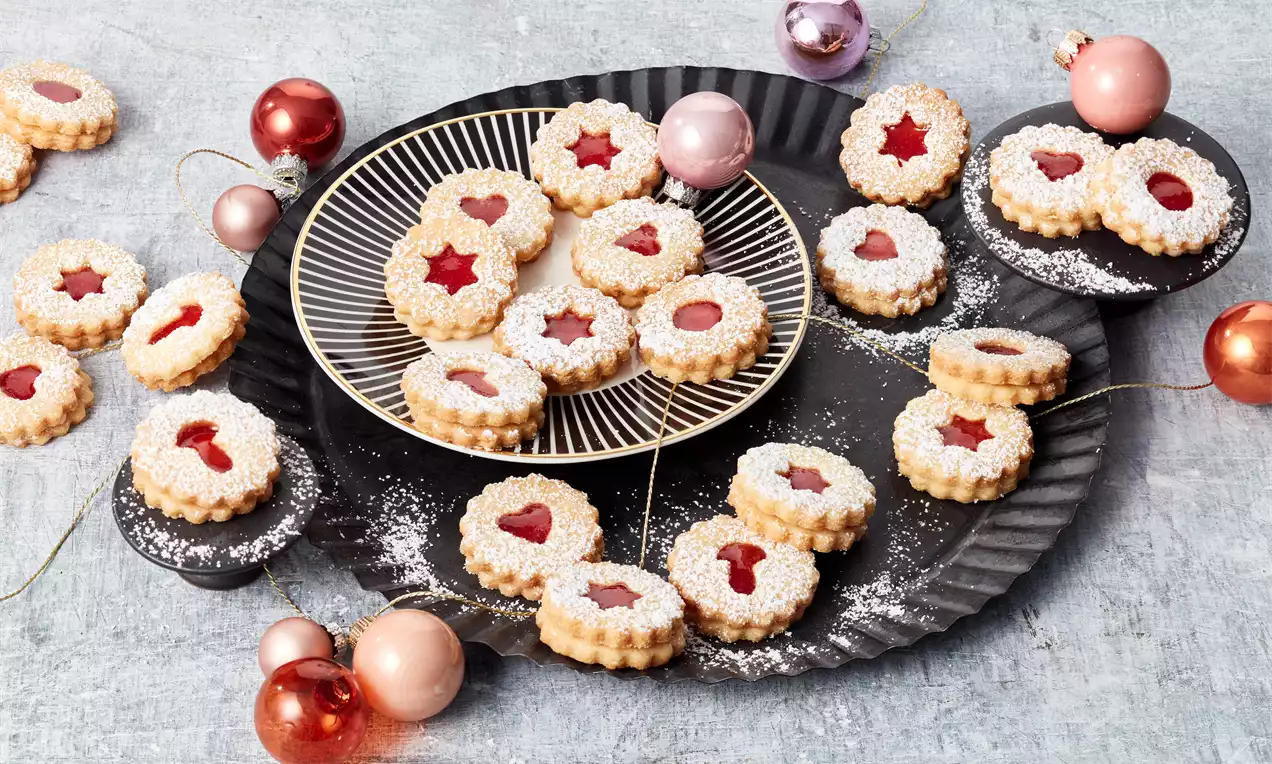 Weihnachts-Spitzbuben mit roter Füllung, gezacktem Rand und Puderzucker auf schwarzen Tellern, dekoriert mit glänzenden Weihnachtskugeln