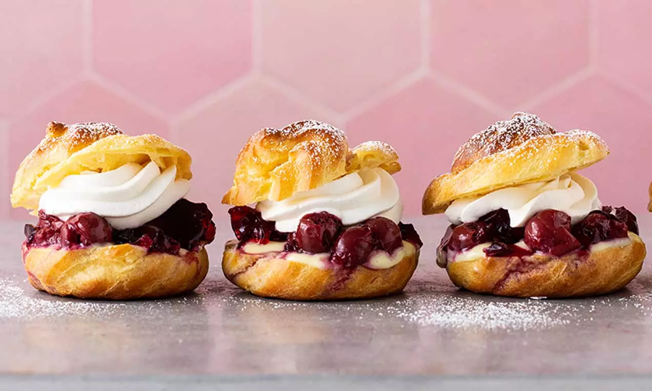 Profiteroles dorati con guscio soffice, ripieni di panna bianca e ciliegie lucide su una base di crema chiara, spolverati di zucchero a velo