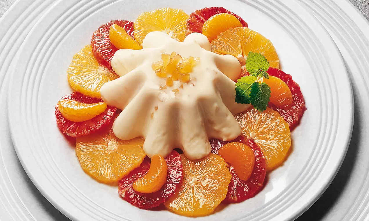 Mousse crémeuse et ondulée couleur ivoire, agrémentée de zestes confits, entourée de tranches d’agrumes vives sur une assiette blanche