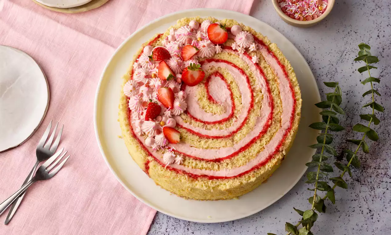 Runde Wickeltorte mit spiralförmigen roten Streifen, hellem Biskuitteig, rosa Creme und frischen Erdbeeren auf weißem Teller mit Dekoration