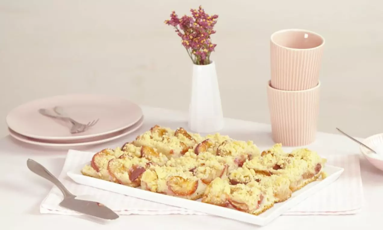 Carrés dorés de gâteau moelleux aux prunes violettes et crumble croustillant, disposés sur un plat blanc avec une décoration douce