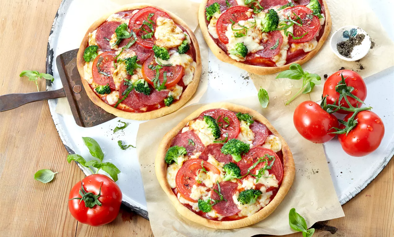 Goldgelbe Mini-Pizza mit lockerem Teig, belegt mit roten Tomatenscheiben, grünen Brokkoliröschen, Salamischeiben und geschmolzenem Käse