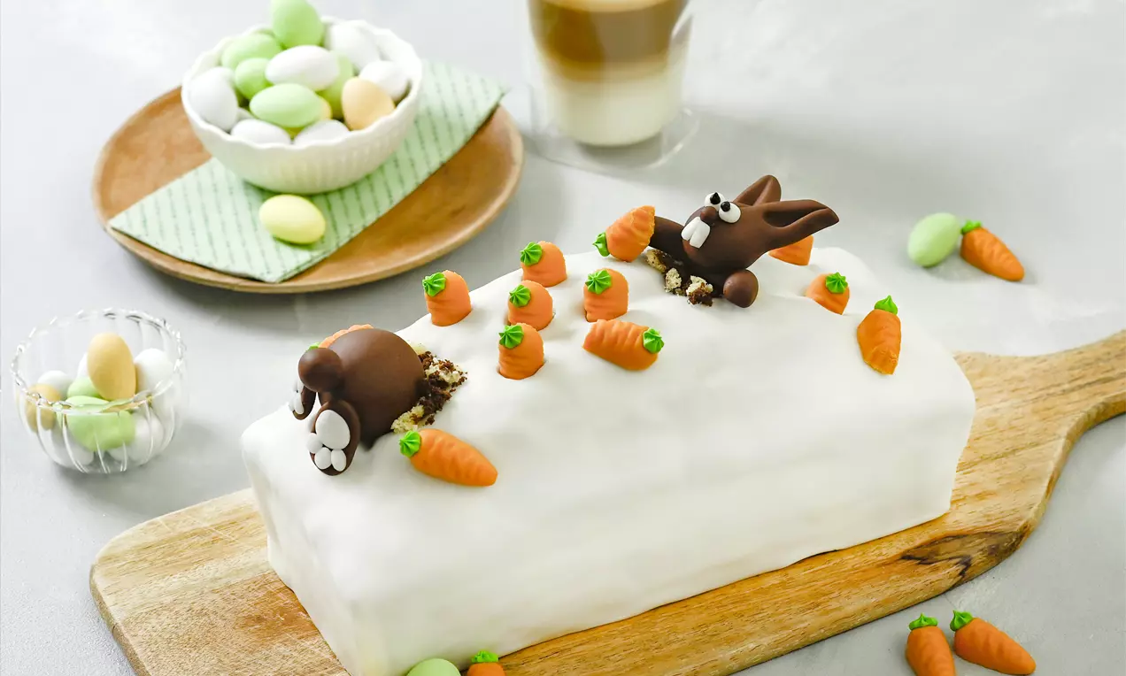 Ein rechteckiger Möhren-Marmorkuchen mit glatter weißer Fondantschicht, verziert mit kleinen orange-grünen Fondantmöhren und braunen Fondanthäschen