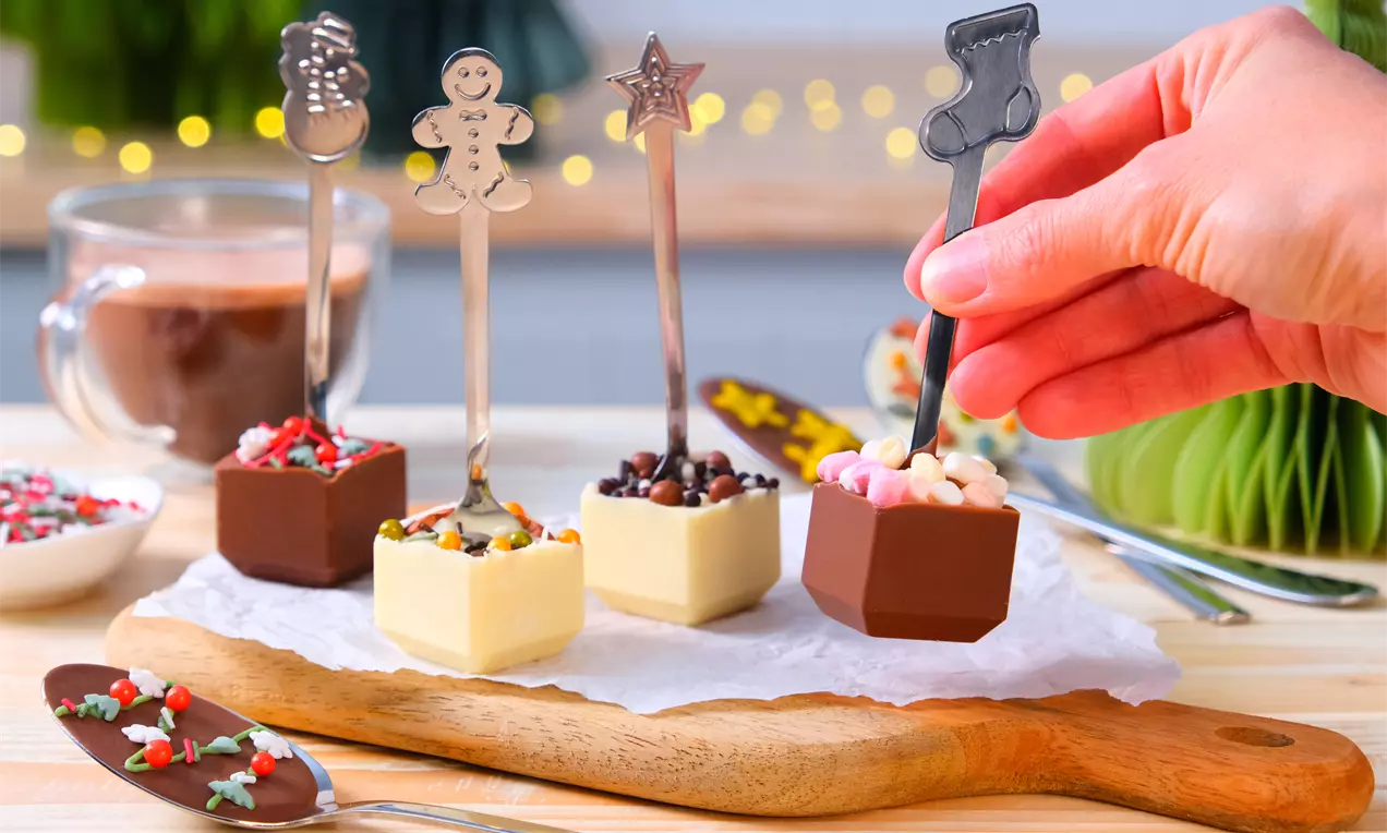 Schokolöffel aus weißer und brauner Schokolade, verziert mit bunten Streuseln und Mini-Marshmallows, angerichtet auf einem Holzbrett mit Dekoration