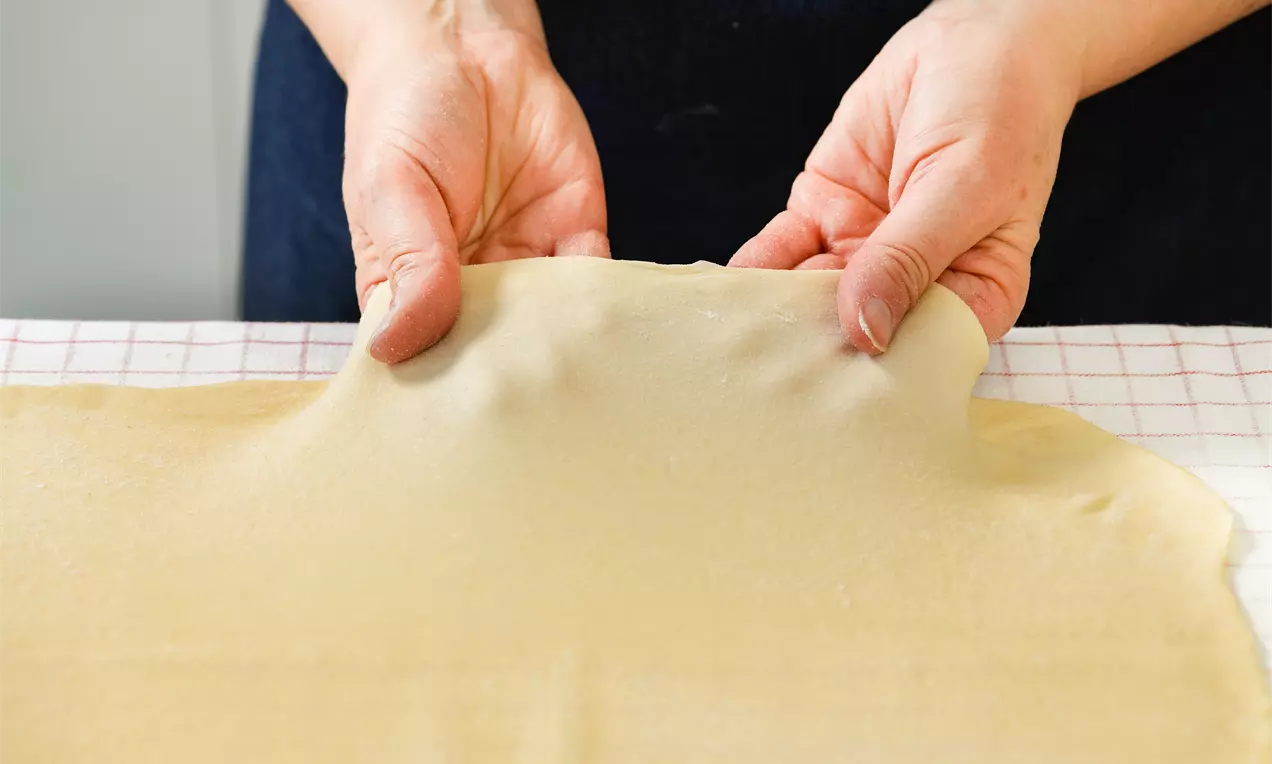 Hände ziehen dünnen, hellgoldenen Strudelteig mit glatter, leicht durchscheinender Oberfläche auf kariertem Tuch behutsam aus