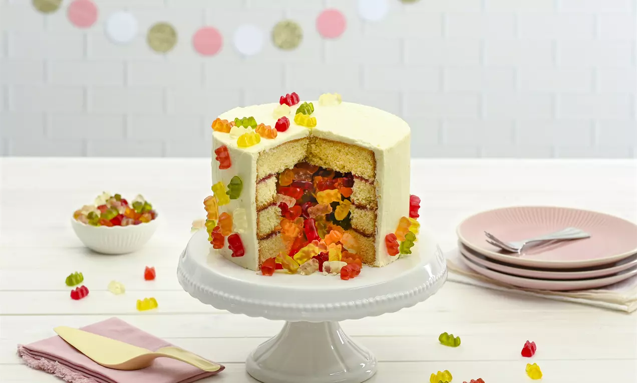 Weiße Cremetorte mit Schichten aus hellem Biskuit, gefüllt mit bunten Goldbären, auf einer weißen Tortenplatte mit rosa Teller dekoriert