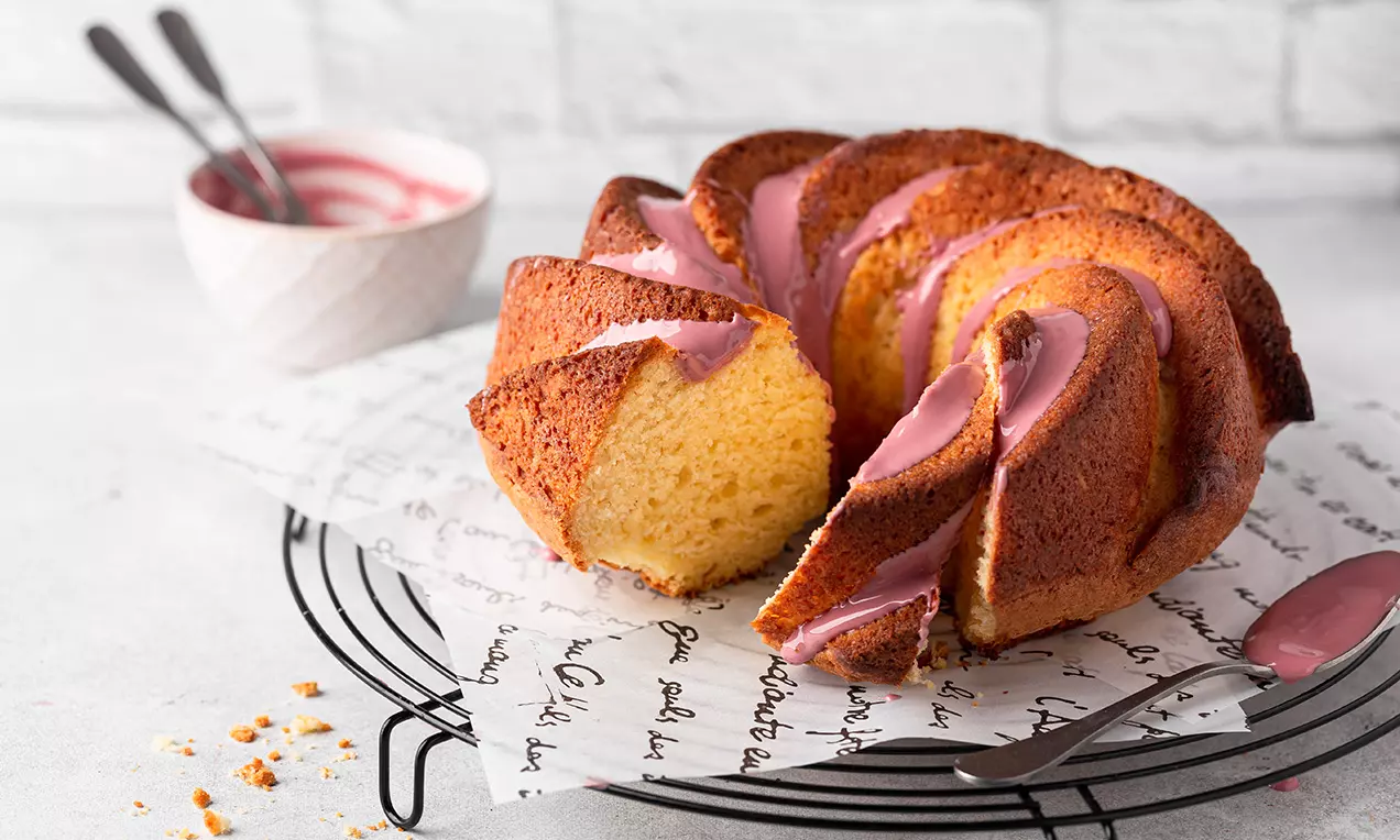 Kouglof moelleux aux tons dorés, glaçage rose délicat coulant le long des courbes, posé sur un papier imprimé sur grille noir