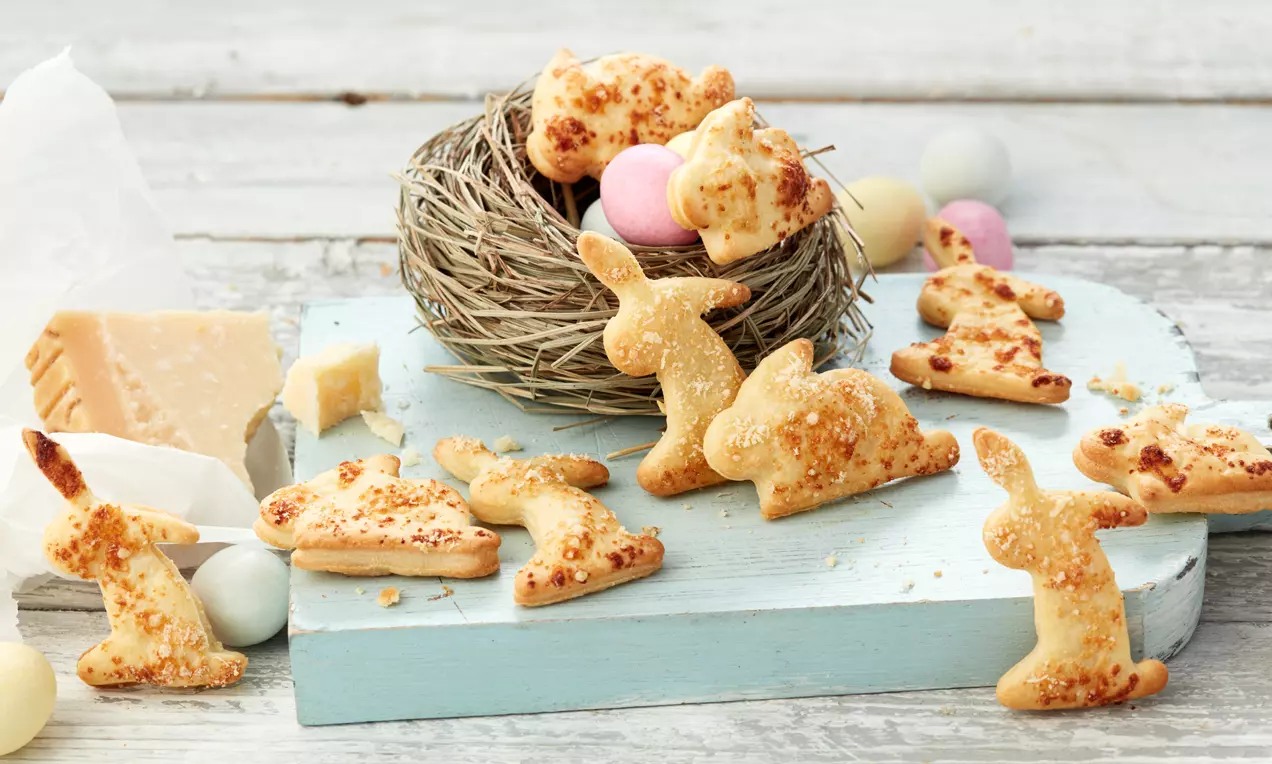 Goldgelbe, knusprige Osterhasen aus Blätterteig mit Käsekruste vor einem dekorativen Nest mit bunten Eiern auf hellblauer Holzplatte
