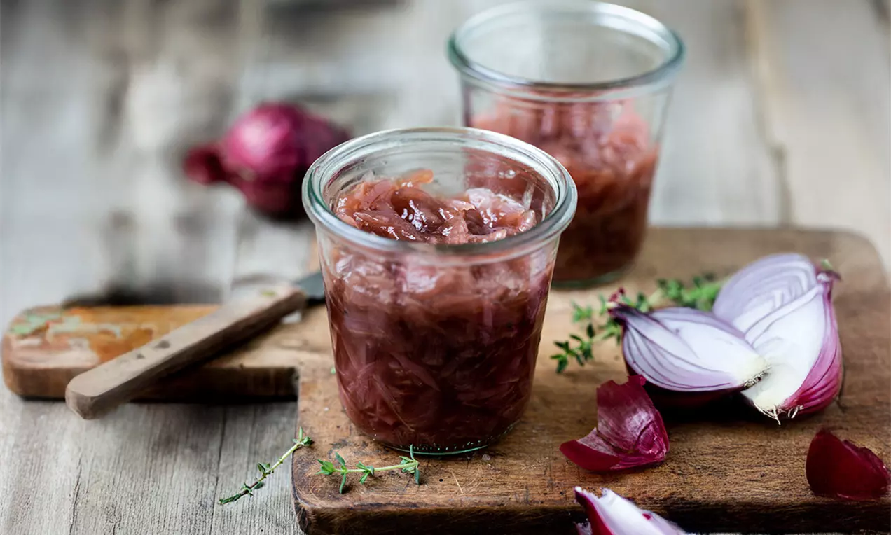 Confit d'oignons rouges à la texture fondante, aux teintes pourpres et translucides, présenté dans un pot de verre sur une planche en bois