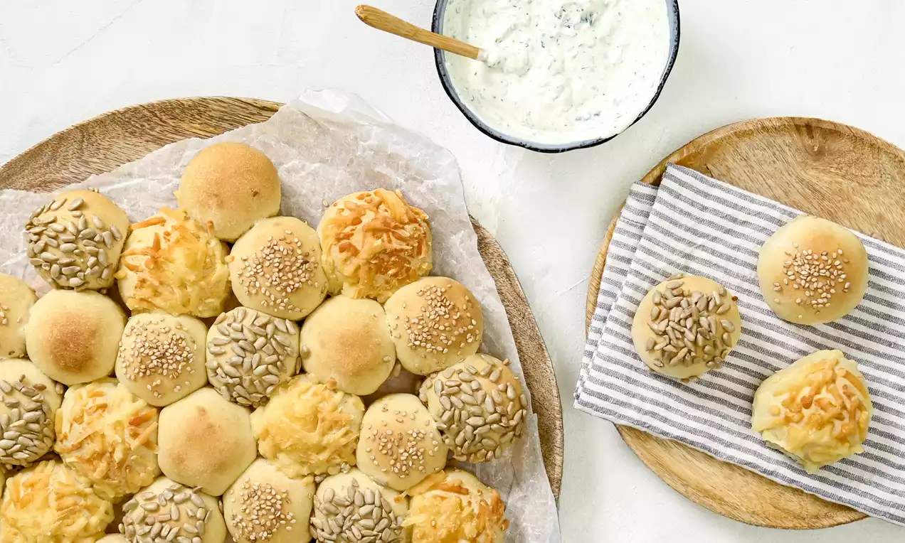 Knoblauchbrot als kleine, goldgelbe Brötchen mit Käse, Sesam und Sonnenblumenkernen auf Holzbrett, daneben Dip und Brotkörbchen