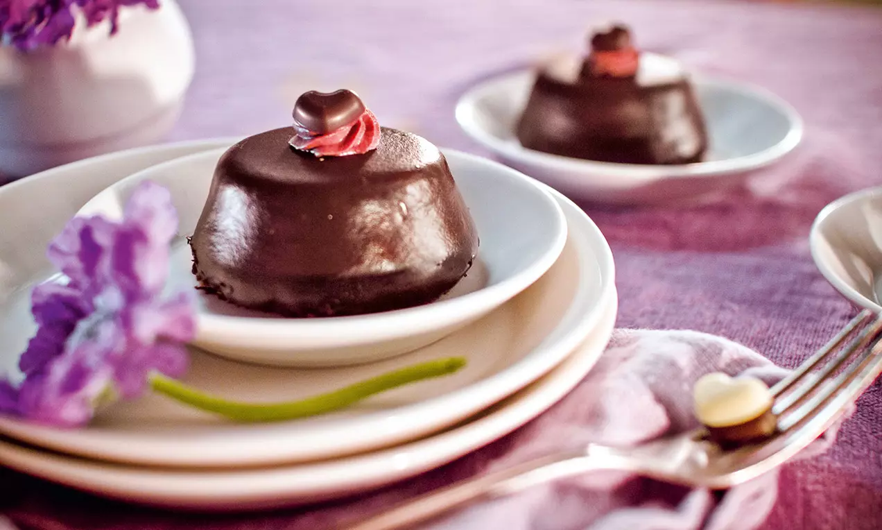 Tortino dal guscio lucido di cioccolato fondente, decorato con crema rossa e cuoricino su piattino bianco, accanto a fiore lilla e posate argento