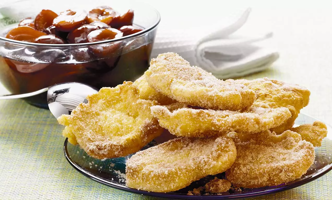 Sfoglie dorate e sottili con bordo irregolare, ricoperte di zucchero scintillante, servite su un piatto accanto a frutta sciroppata in ciotola di vetro