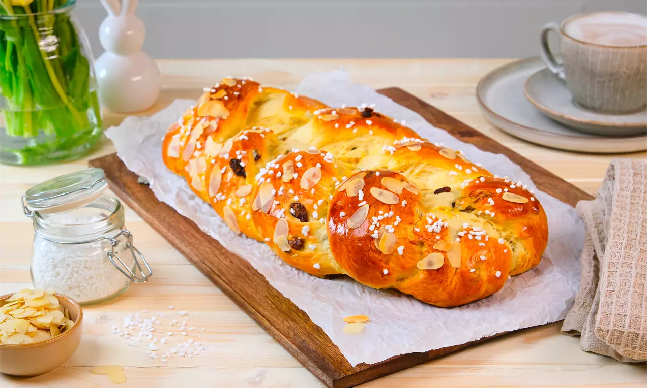 Brioche dorée tressée en trois brins, parsemée d’amandes effilées et de sucre perlé, posée sur du papier parchemin sur une planche en bois