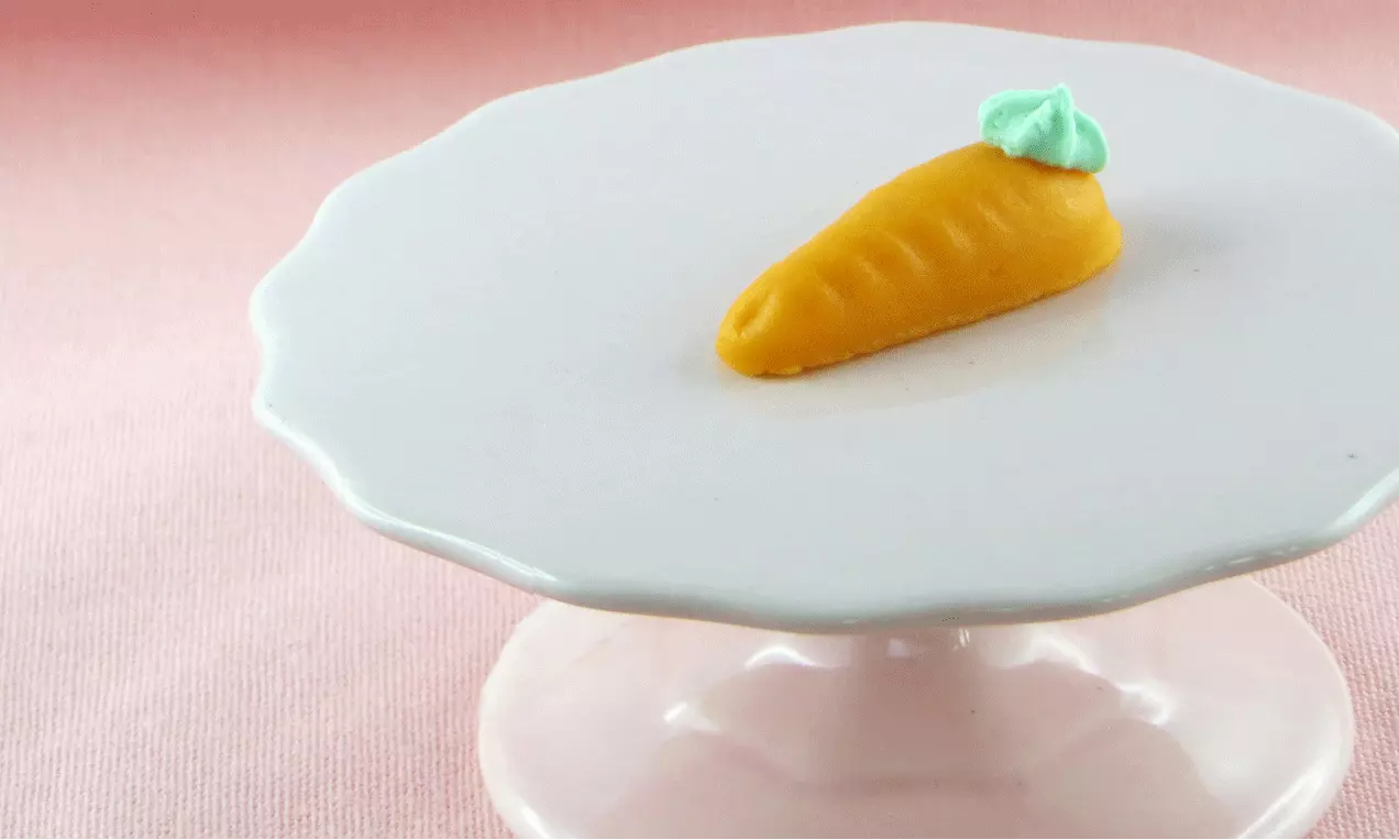 Petite carotte en pâte de massepain orange avec une feuille verte en relief, posée sur un support rond blanc aux bords ondulés