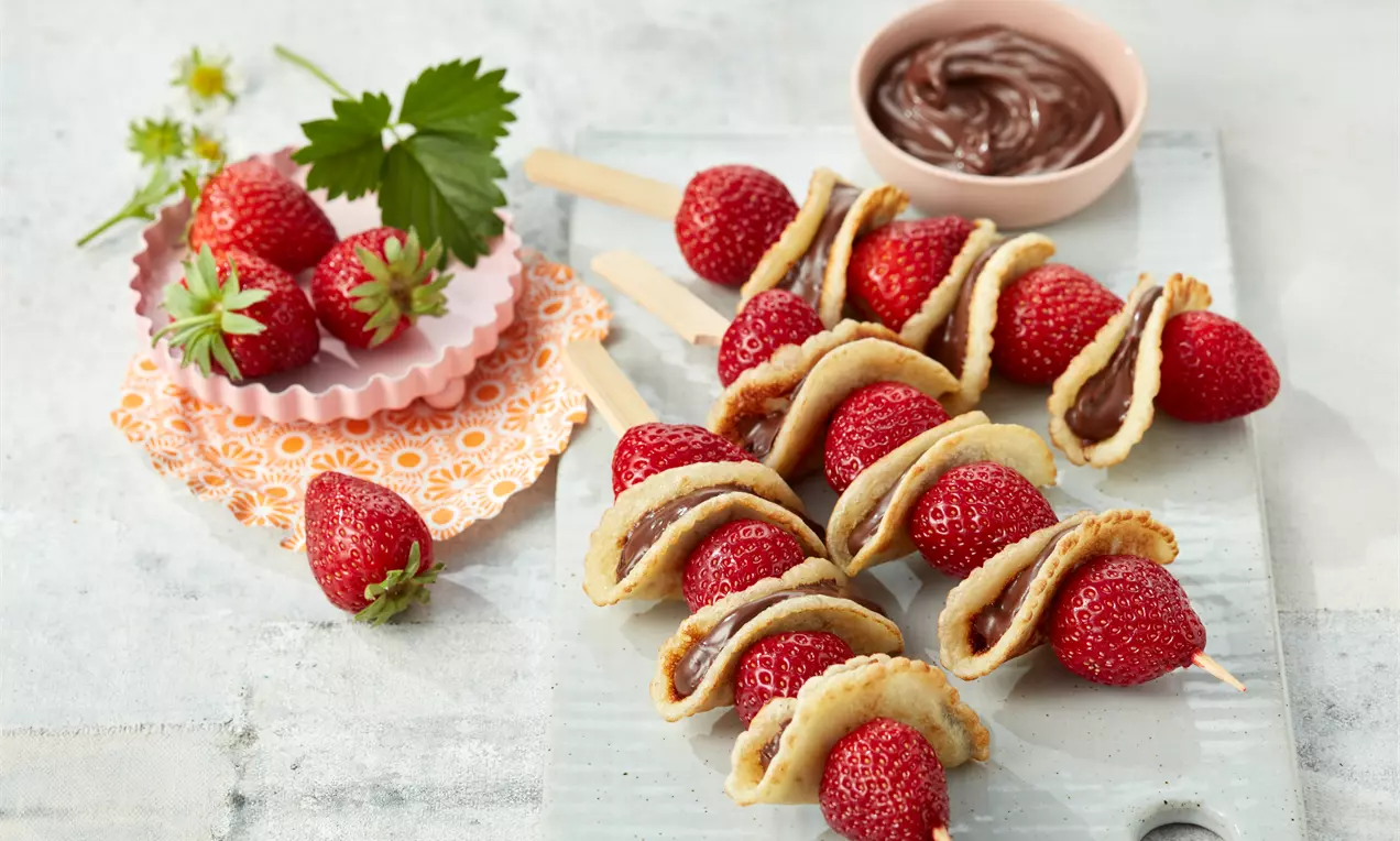 Goldbraun gebackene Püfferchen abwechselnd mit saftigen roten Erdbeeren auf Holzspießen, serviert auf hellem Untergrund mit Schokocreme im Hintergrund