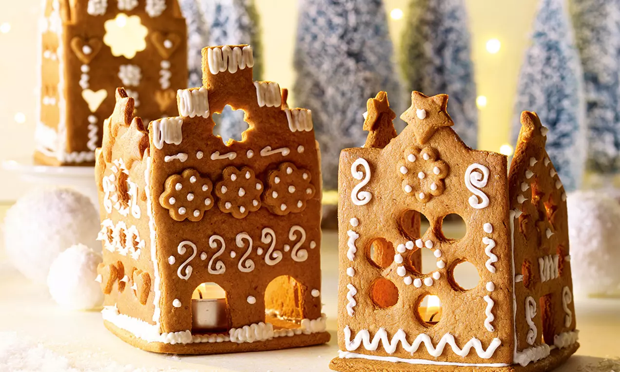 Zwei goldbraune Lebkuchenhäuschen mit weißem Zuckerguss verziert, innen beleuchtet vor schneebestäubten Miniaturtannen im Hintergrund
